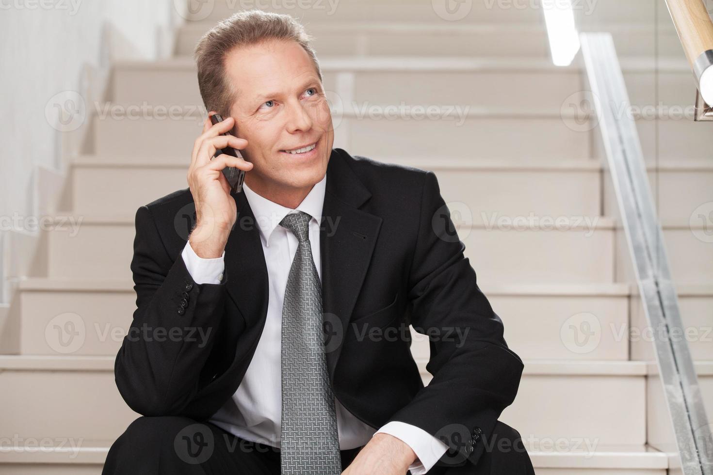 He is always in touch. Cheerful mature man in formalwear talking on the mobile phone and smiling while sitting on staircase photo