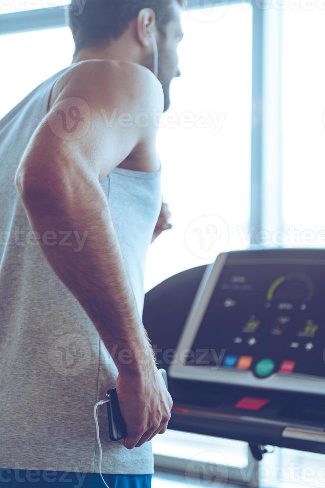 Exercising with great music. Rear view of young man in headphones running on treadmill in front of window at gym and holding his smart phone in his hand photo