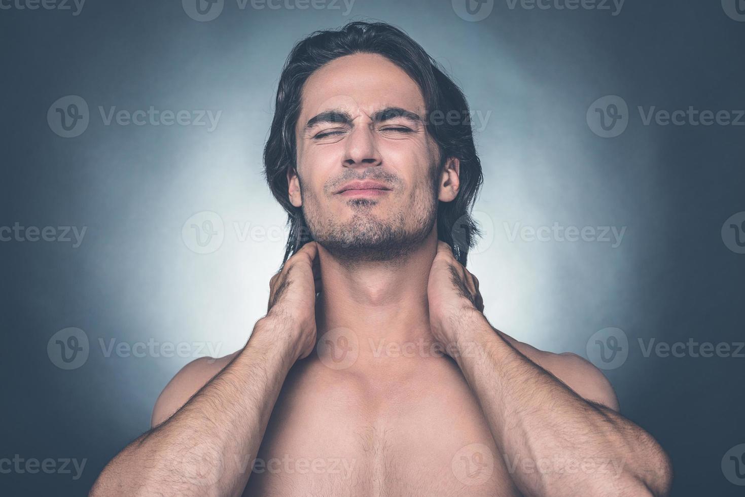 Feeling that awful pain. Portrait of young shirtless man keeping eyes closed and expressing negativity while touching his neck and standing against grey background photo