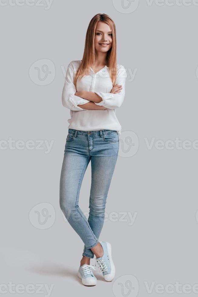 Confident woman. Full length of beautiful young woman in casual wear keeping arms crossed and looking at camera with smile while standing against grey background photo