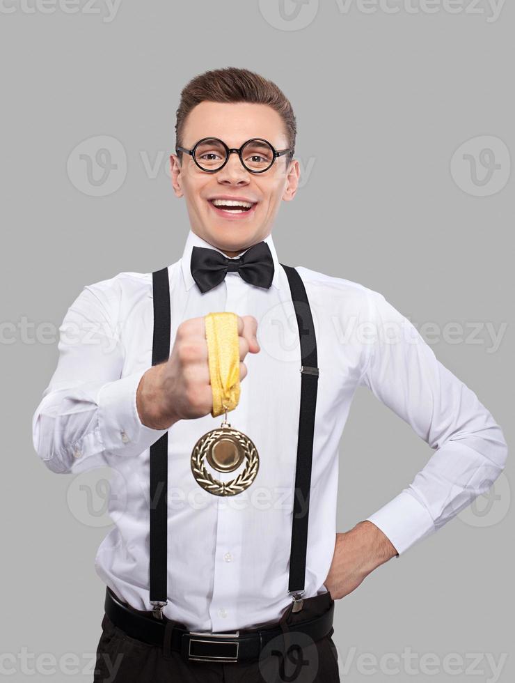 I am winner Cheerful young man in bow tie and suspenders showing a gold medal to camera and smiling while standing against grey background photo