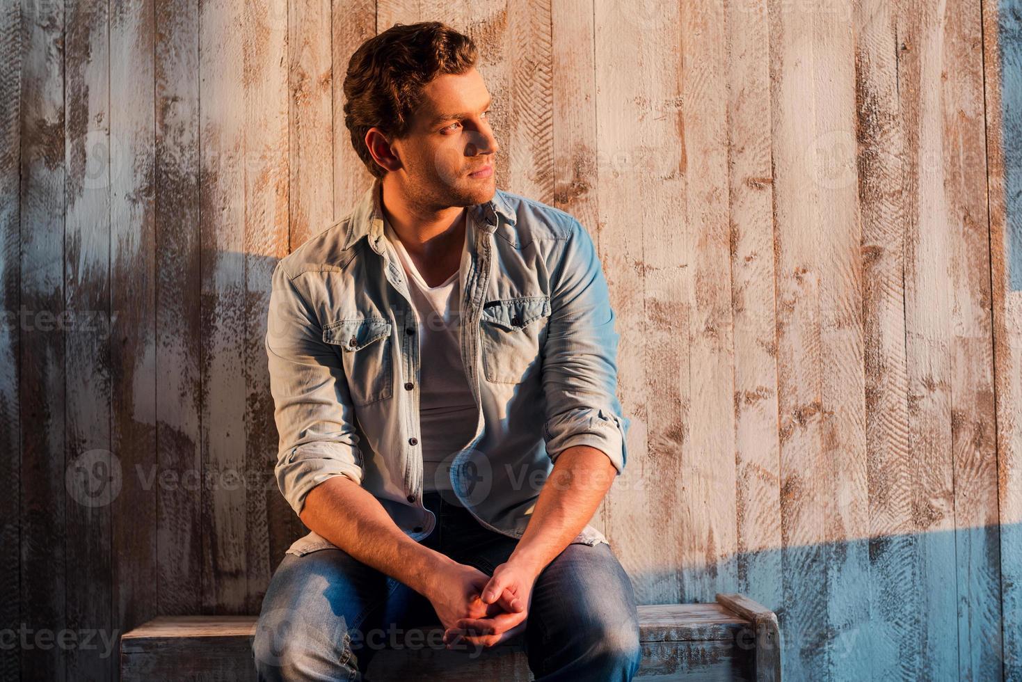 encantador guapo. retrato de un apuesto joven mirando hacia otro lado mientras se sienta contra la pared de madera foto