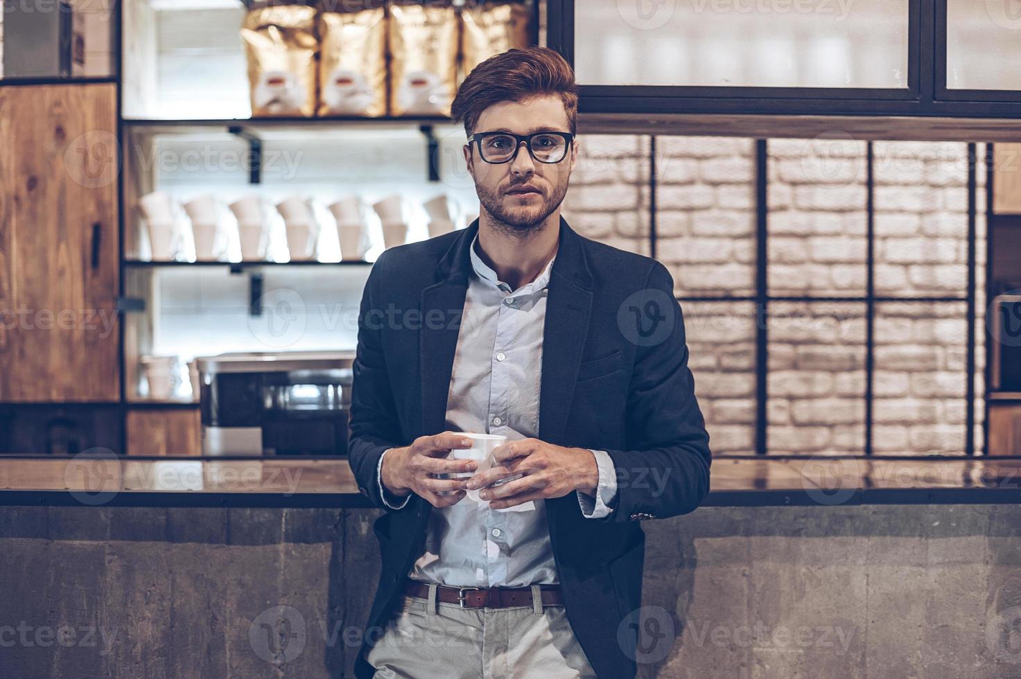 iniciando su pequeña empresa. joven apuesto con gafas sosteniendo una taza de café y mirando a la cámara mientras está de pie en el mostrador del bar foto