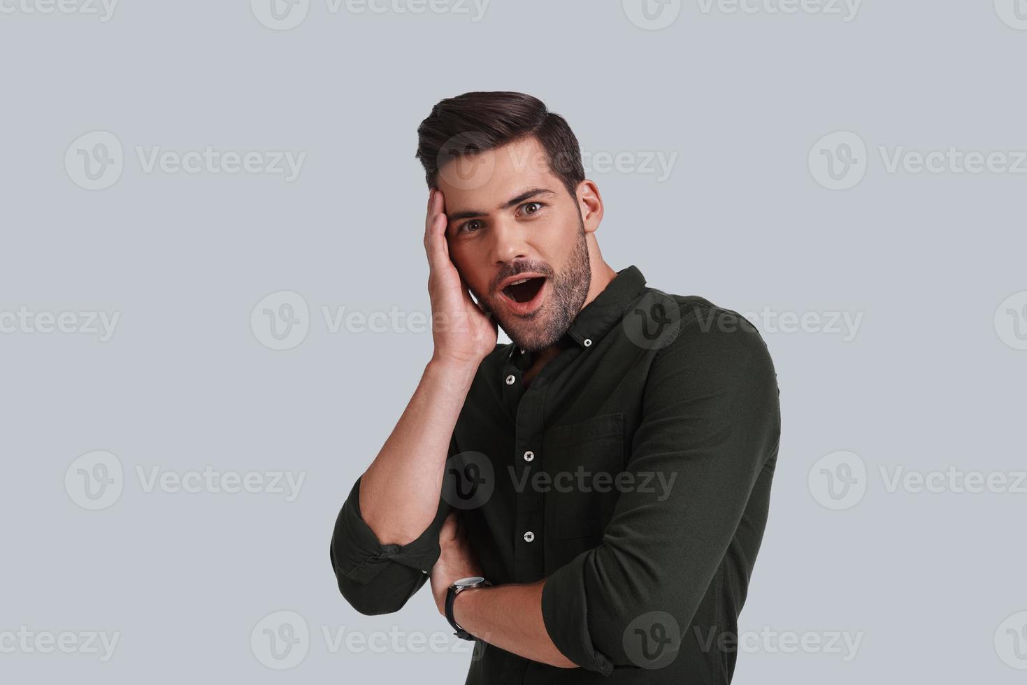 Feeling surprised. Shocked young man keeping mouth open and looking at camera while standing against grey background photo