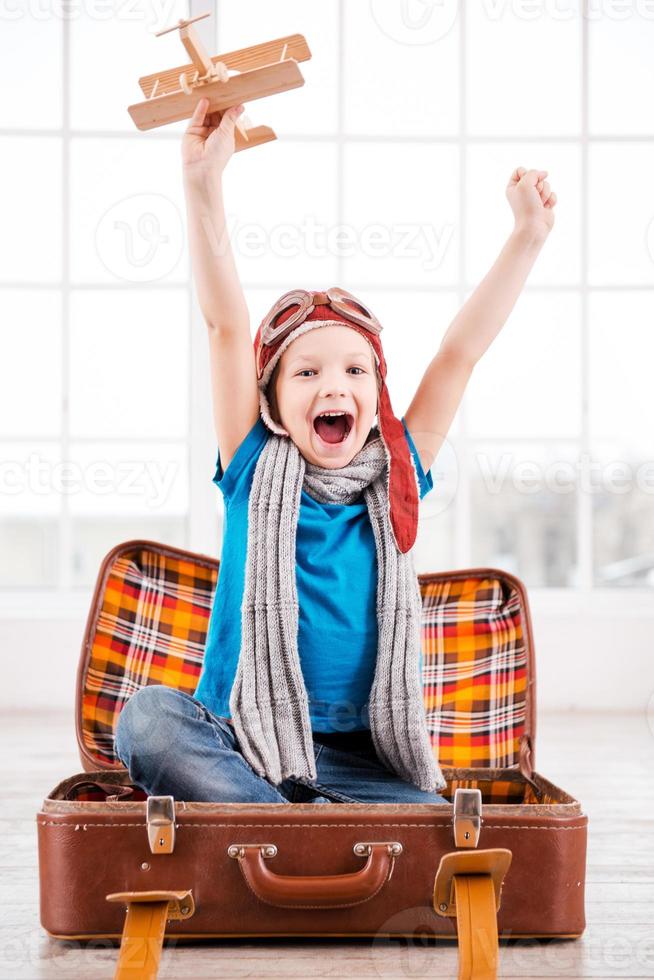 Little pilot. Happy little boy in pilot headwear and eyeglasses playing with wooden planer and smiling while sitting inside of briefcase at home photo