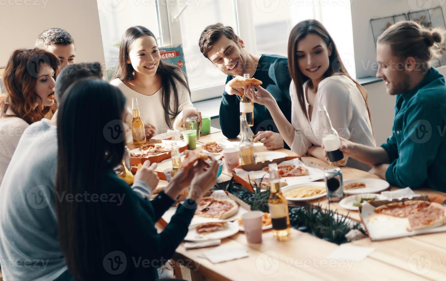 fiesta inolvidable. grupo de jóvenes con ropa informal comiendo pizza y sonriendo mientras cenan en el interior foto