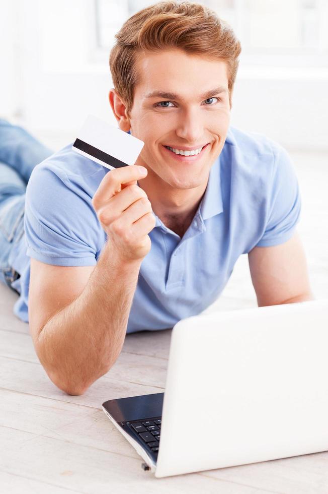 Home shopping. Handsome young man working on laptop and holding credit card while lying on the floor at his apartment photo