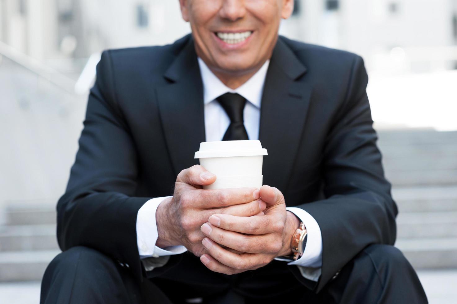 Relaxing with cup of coffee. Cropped image of cheerful mature man in formalwear holding coffee cup and smiling while sitting at the staircase outdoors photo