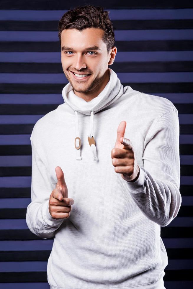 Cool handsome. Handsome young man in white sweater looking at camera and pointing you with smile while standing against striped background photo