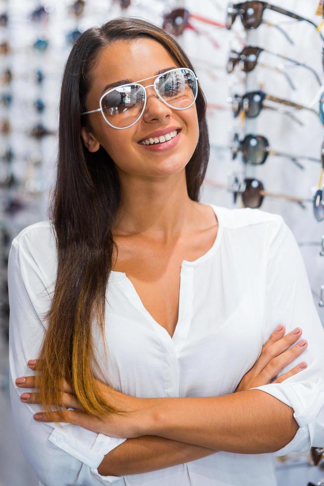 I made my choice. Beautiful young woman in sunglasses keeping arms crossed and smiling while standing in optic store photo