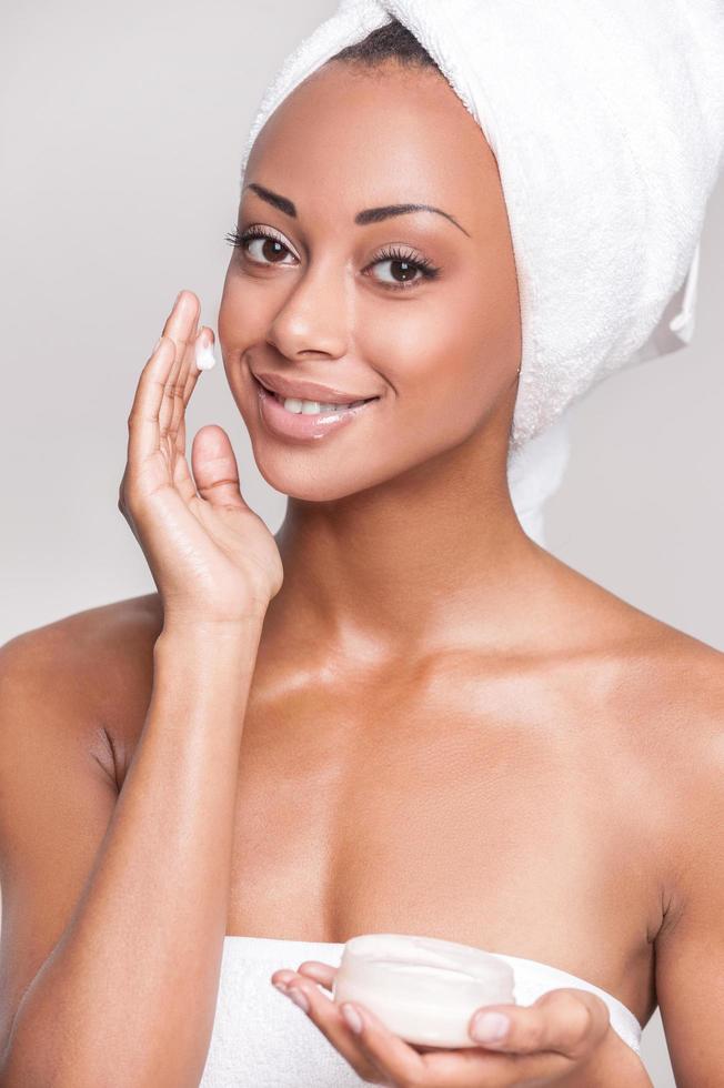 Taking care of her skin. Portrait of beautiful young Afro-American woman wrapped in towel spreading cream on her face and smiling while Isolated on gray background photo
