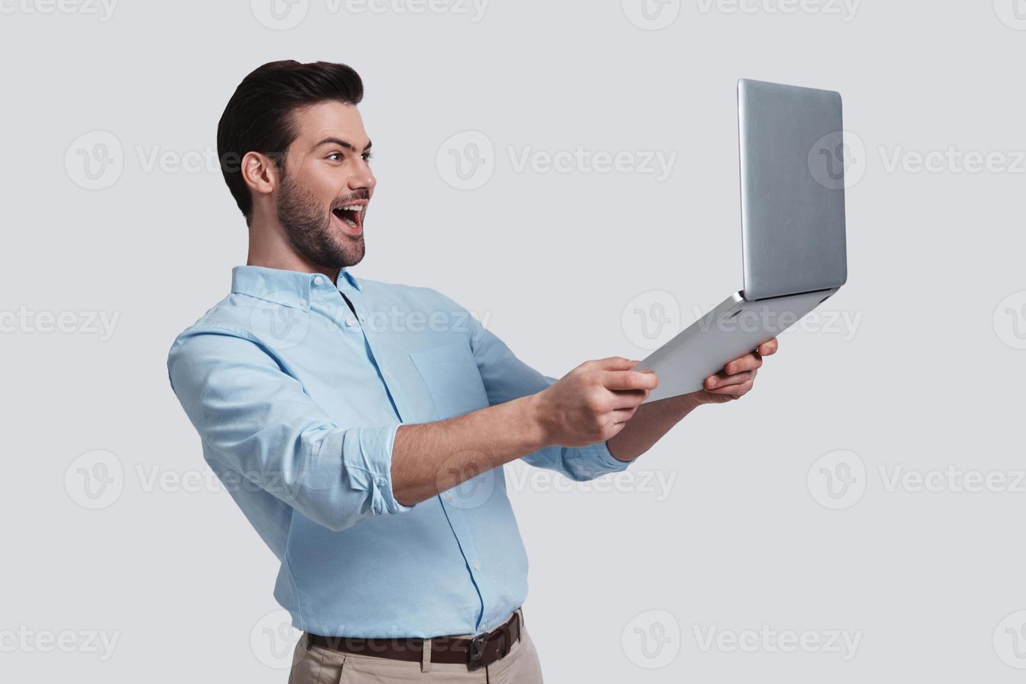 Feeling happy.  Good looking young man holding his laptop and smiling while standing against grey background photo