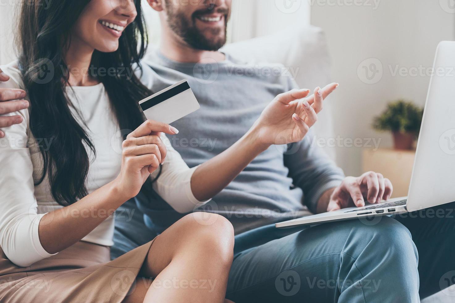 quiero este primer plano de una hermosa joven sosteniendo una tarjeta de crédito y señalando una laptop con una sonrisa mientras se sienta junto a su esposo en el sofá foto