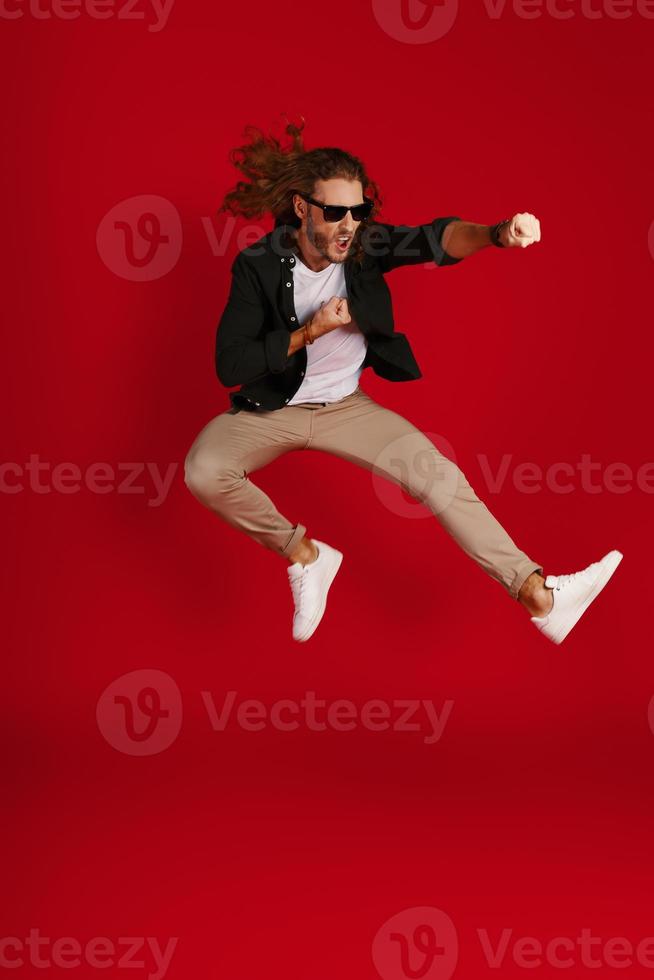 Full length of playful young man in casual clothing making a face and punching while hovering against red background photo
