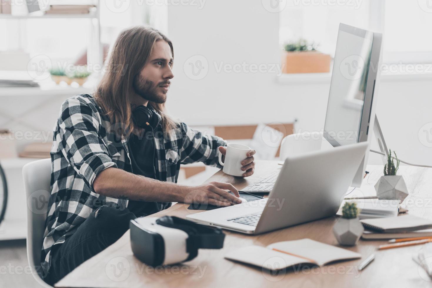 Achieving best results. Handsome young man with long hair working on the computer and holding a cup of coffee while sitting at his desk in creative office photo