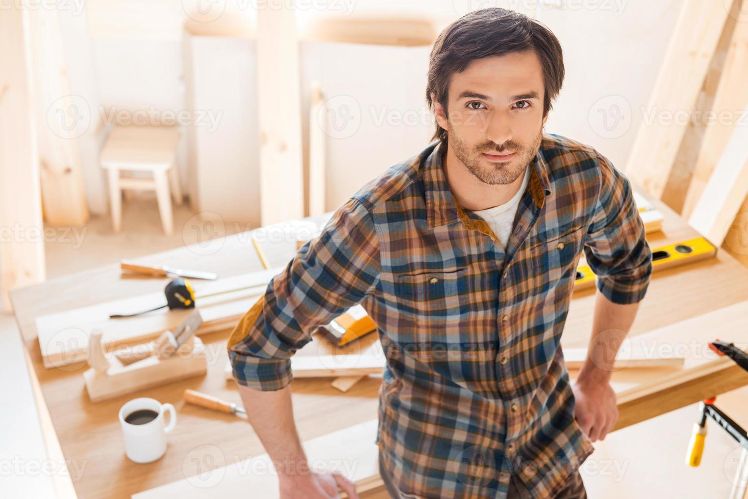 Master of his trade. Top View of confident young male carpenter leaning at the wooden table with diverse working tools laying on it photo
