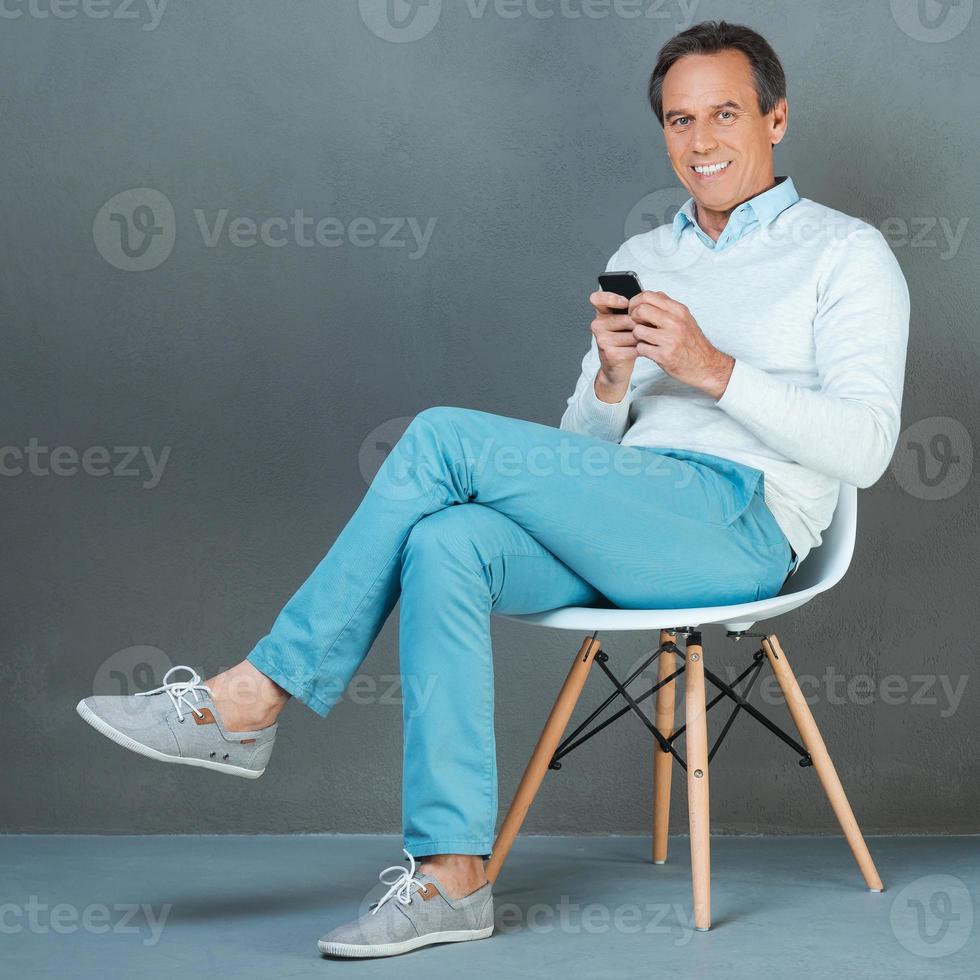 confianza y experiencia. hombre maduro feliz sosteniendo el teléfono móvil y sonriendo a la cámara mientras se sienta en la silla contra el fondo gris foto