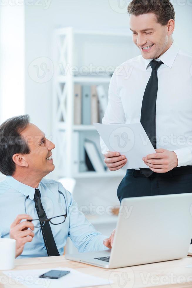 Discussing contract. Smiling mature businessman sitting at his working place while young man standing near with documents photo