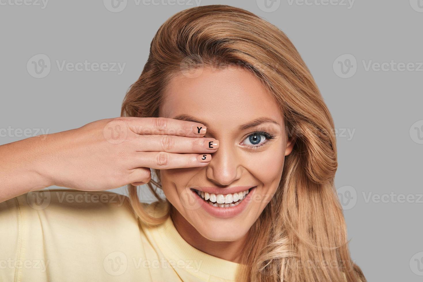 Attractive young smiling woman covering eye with hand and looking at camera while standing against grey background photo
