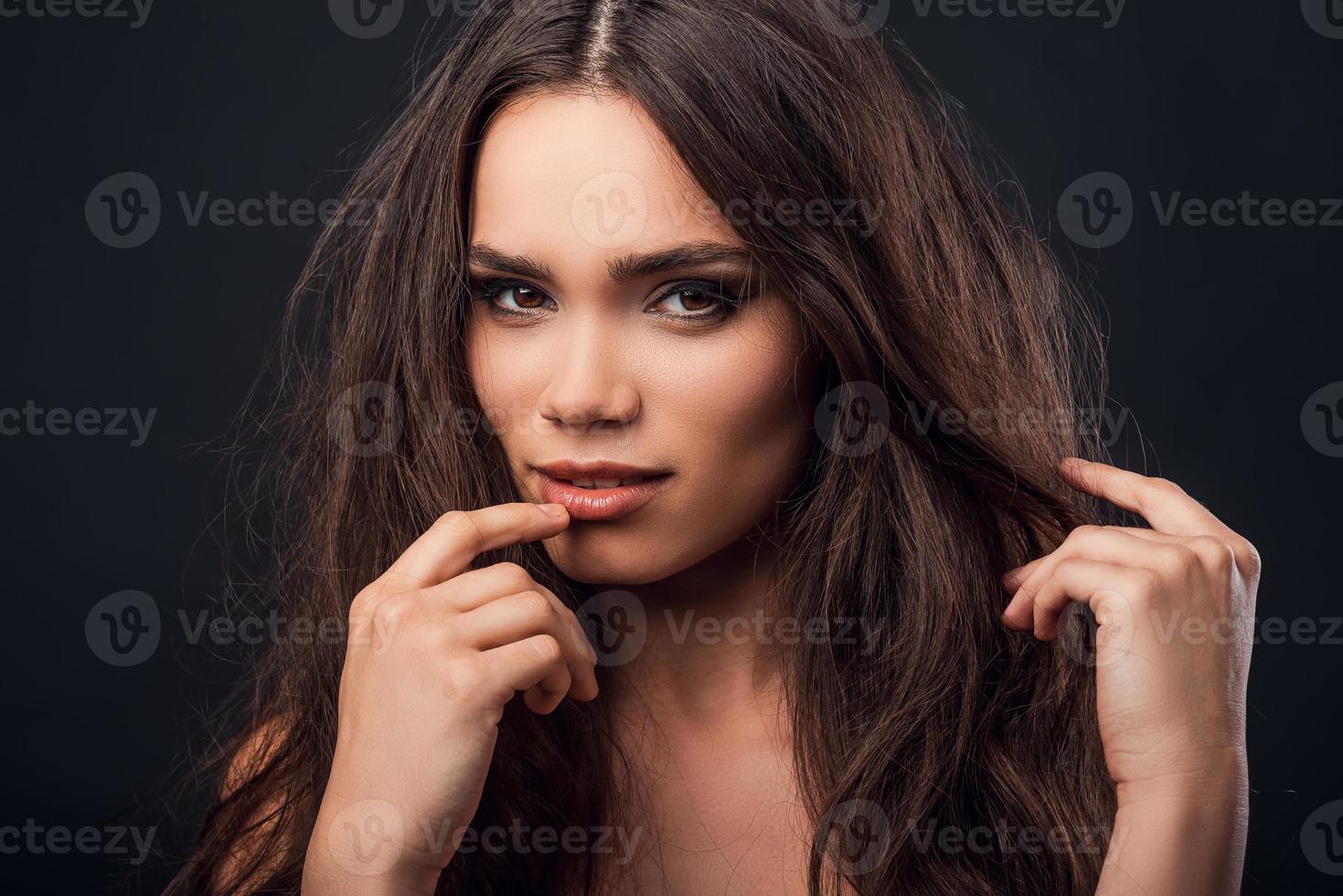 Young and flirty. Beautiful young shirtless woman touching her lips and looking at camera while standing against black background photo