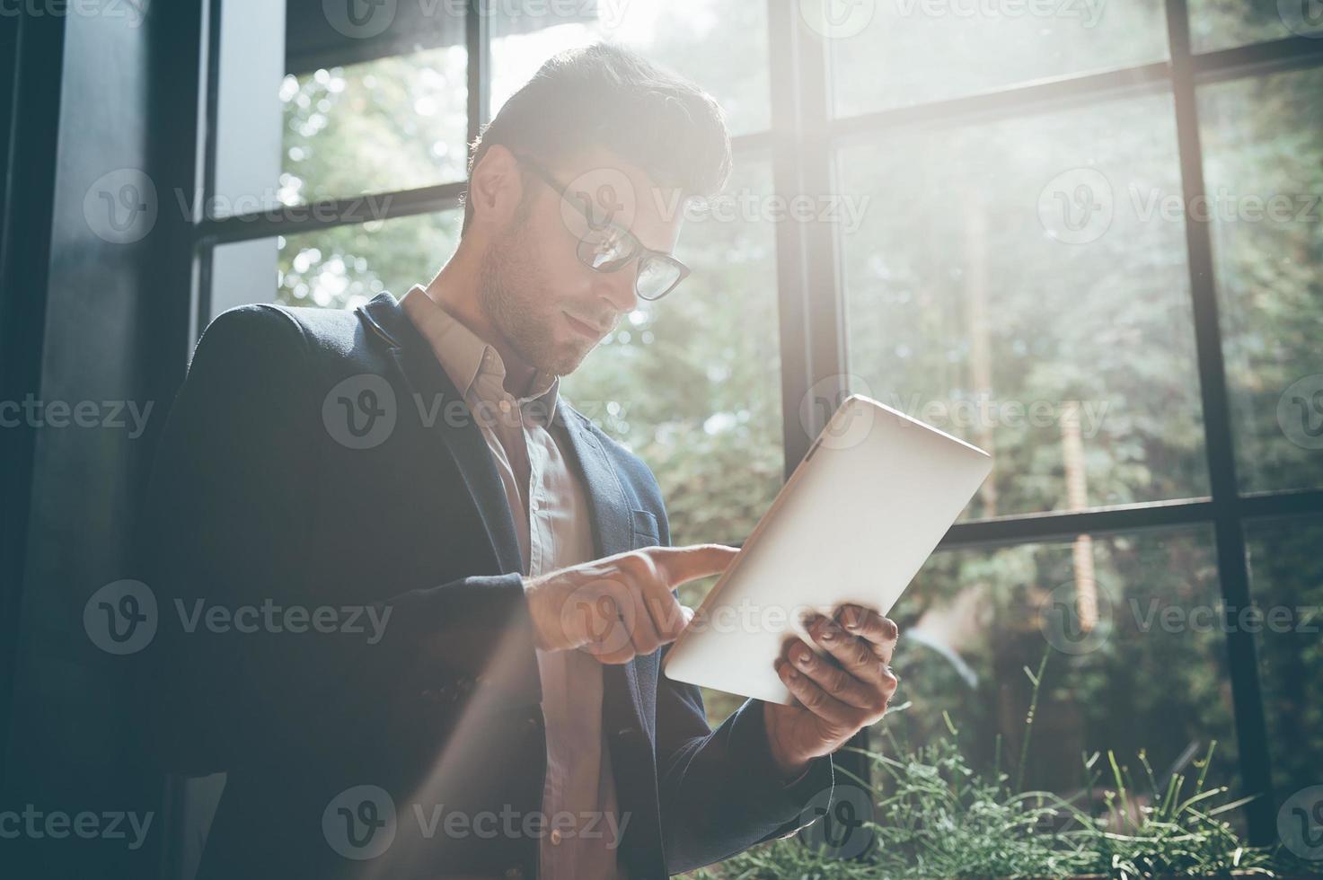 Concentrated on his new project. Low angle view of young man working on digital tablet while standing in front of the big window indoors photo
