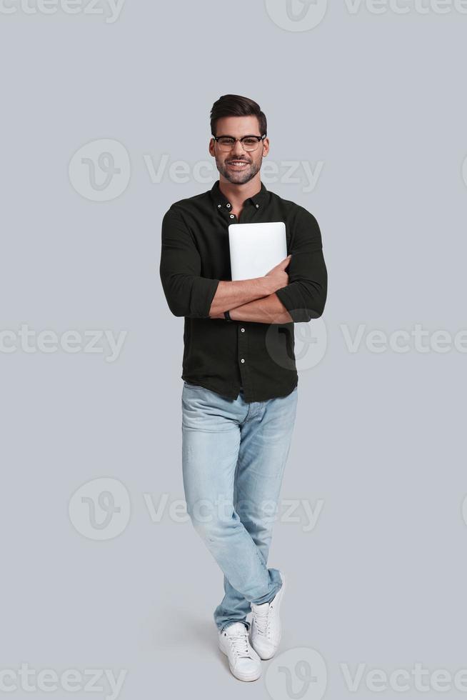 More than just device. Full length of good looking young man in eyeglasses holding his digital tablet and smiling while standing against grey background photo