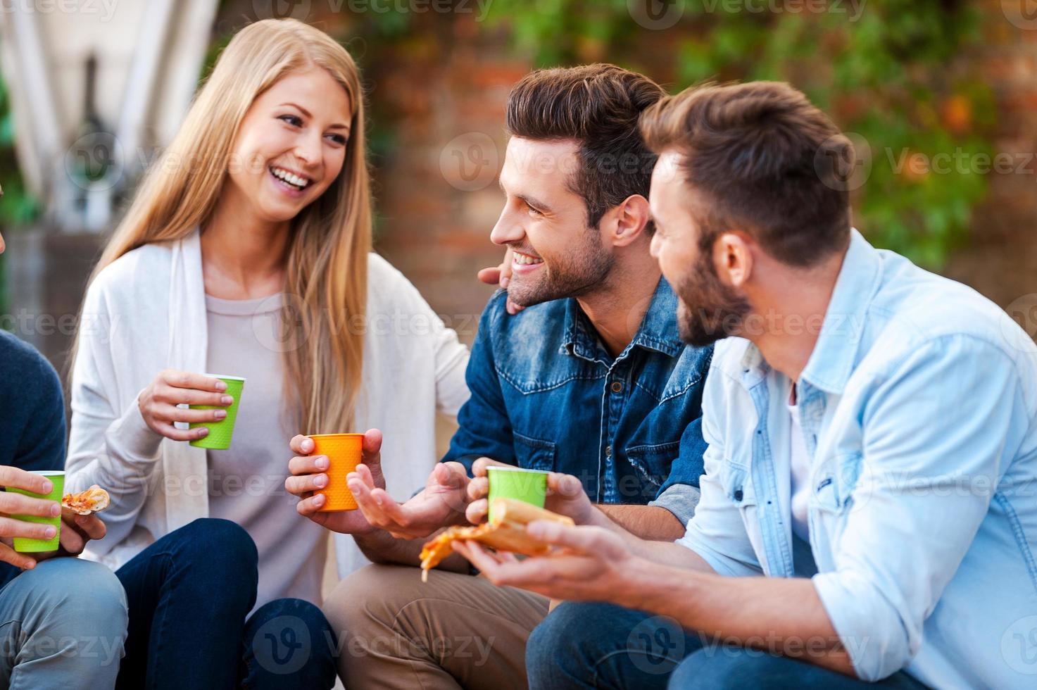 Quality time with best friends. Group of happy young people talking to each other and eating pizza while sitting outdoors photo