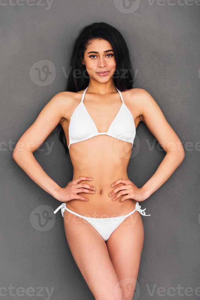 Looking gorgeous. Beautiful young mixed race woman in white bikini holding hands on hip and smiling while standing against grey background photo