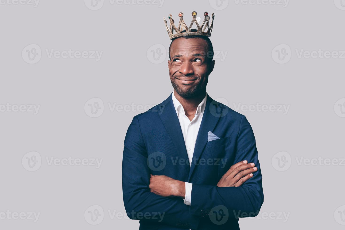 rey apuesto joven africano con corona y elegante chaqueta informal haciendo una mueca mientras está de pie contra un fondo gris foto