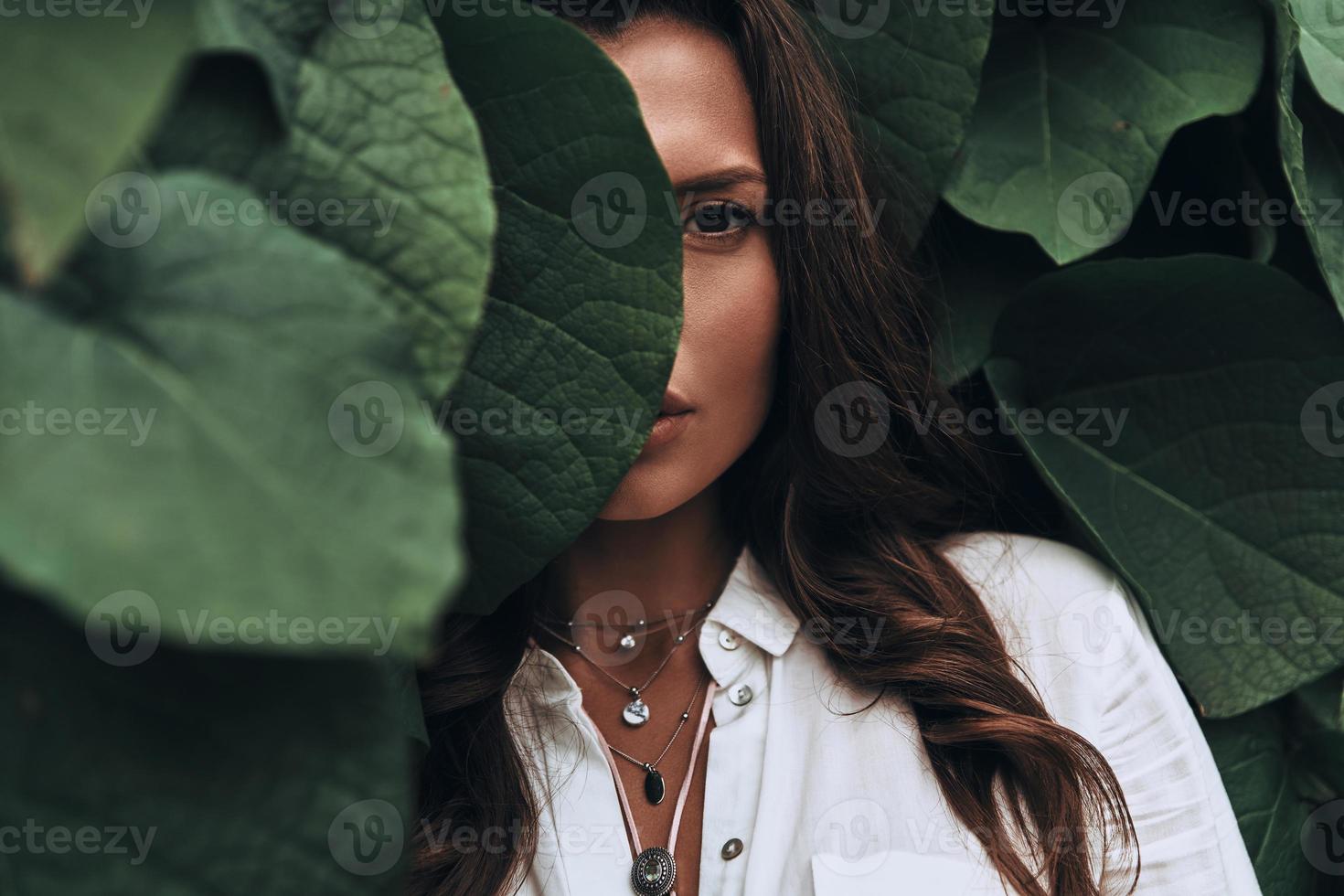 naturalmente bello. primer plano de mujer joven mirando a la cámara mientras está de pie entre las hojas al aire libre foto