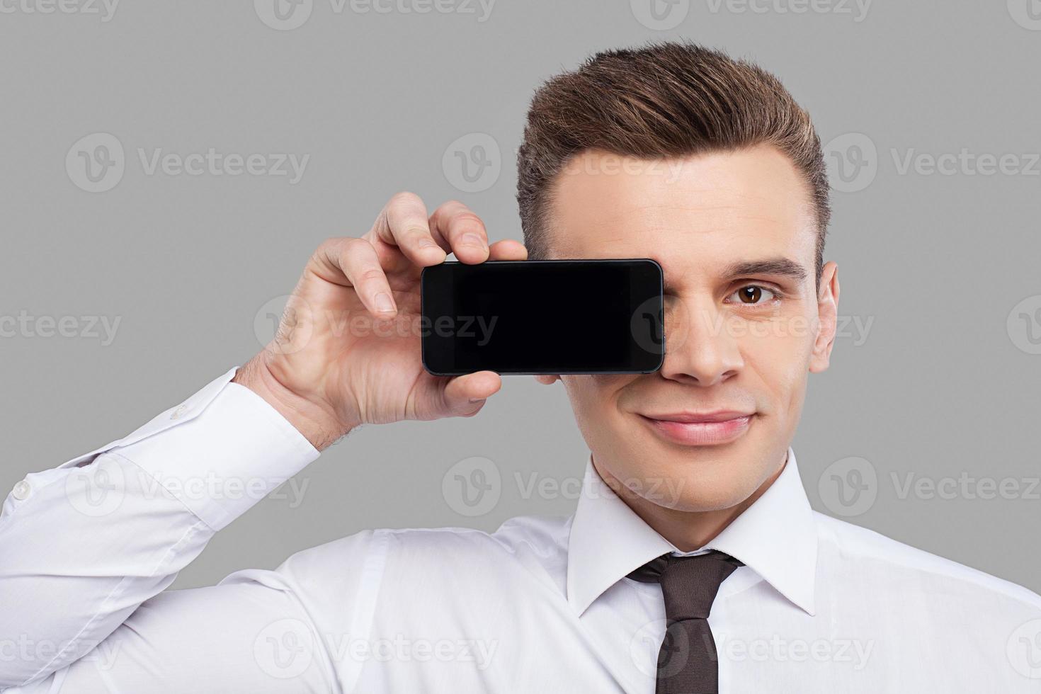 Man with mobile. Handsome young man in shirt and tie holding mobile phone in front of the eye and smiling while standing against grey background photo