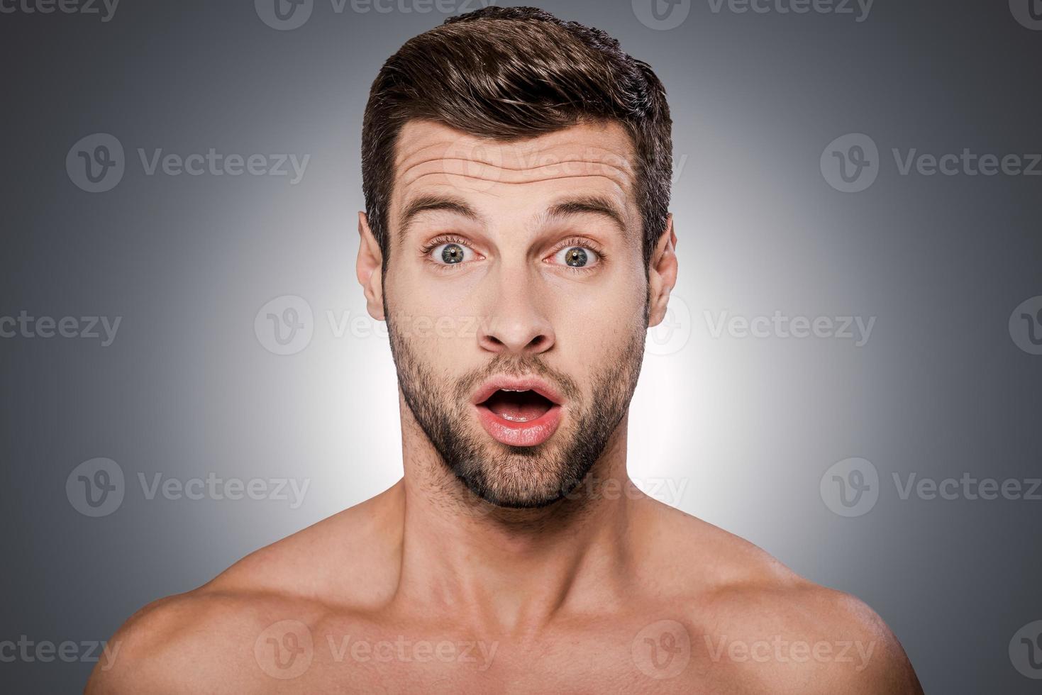 Unbelievable Portrait of surprised young shirtless man looking at camera and keeping mouth open while standing against grey background photo