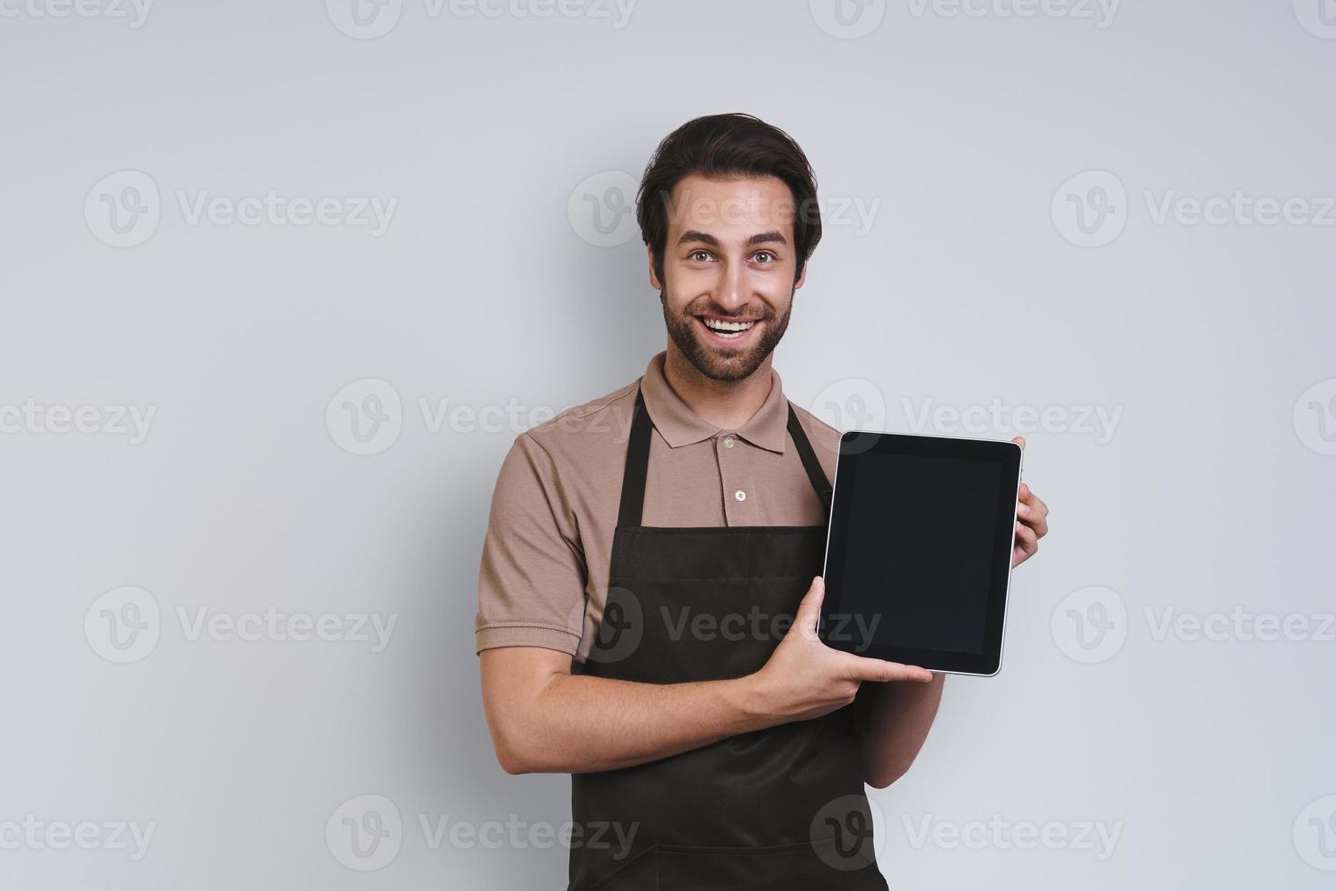 Cheerful young man in apron showing his digital tablet and smiling while standing against gray background photo