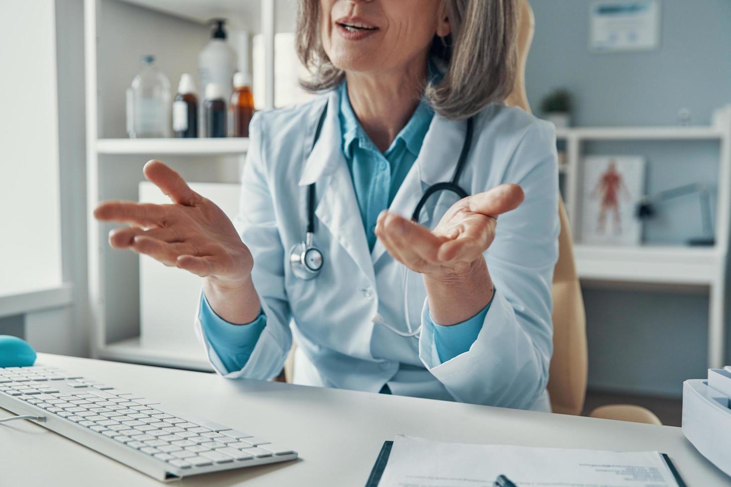 Close up of mature female doctor in white lab coat telling something and gesturing while sitting in her office photo