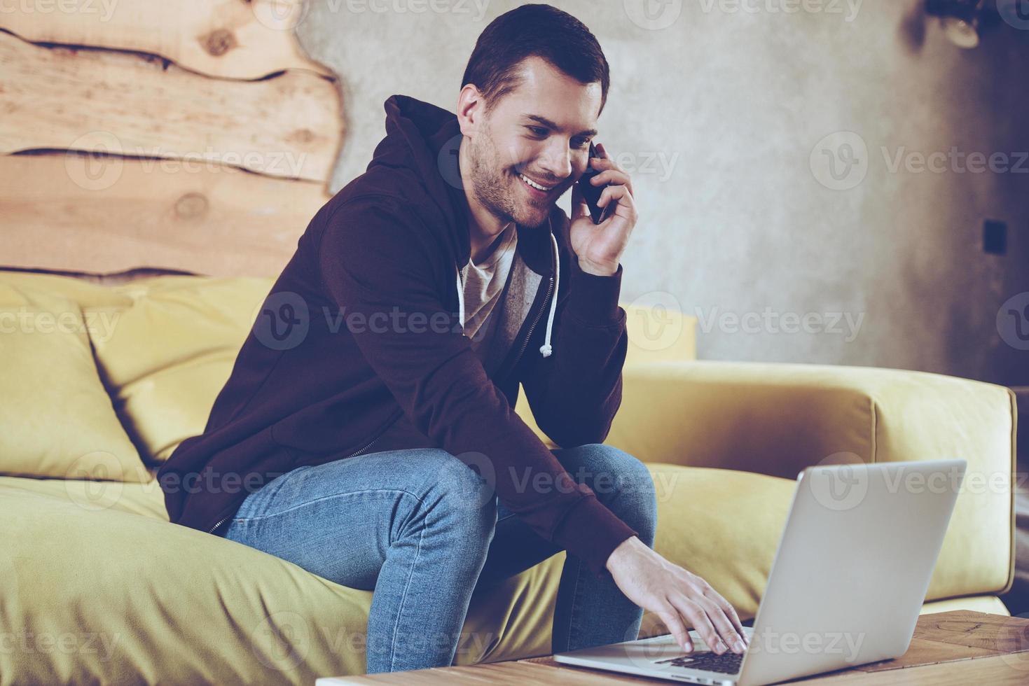 trabajando desde casa con gusto. joven alegre usando su laptop y hablando por teléfono móvil con una sonrisa mientras se sienta en el sofá en casa foto