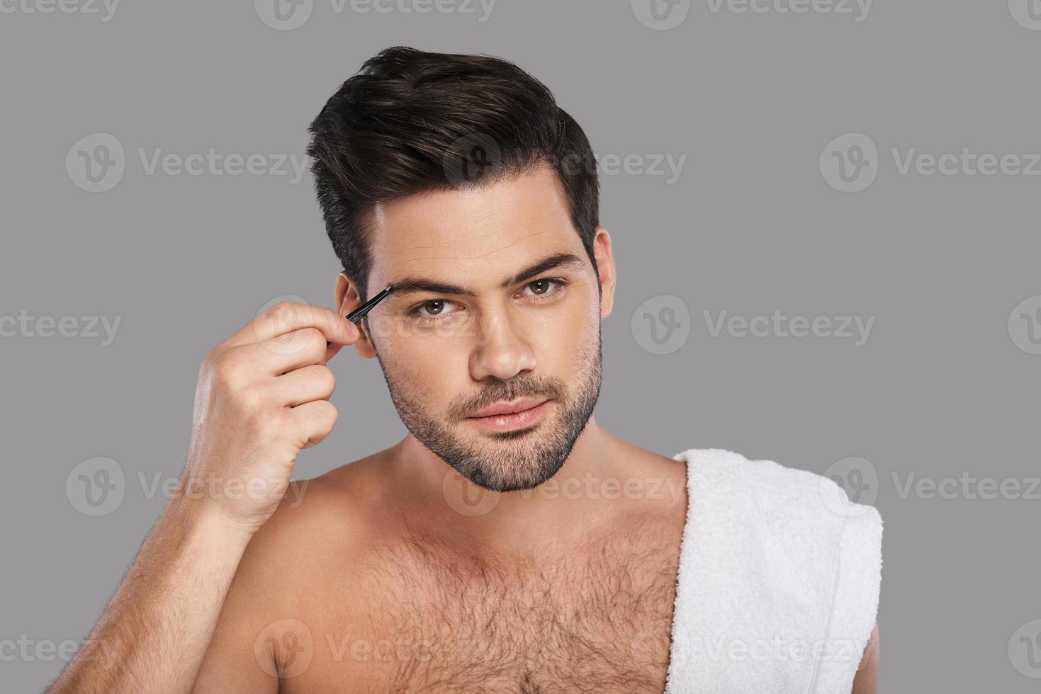 Another beauty treatment. Handsome young man looking at camera and applying eyebrow tweezers while standing against grey background photo