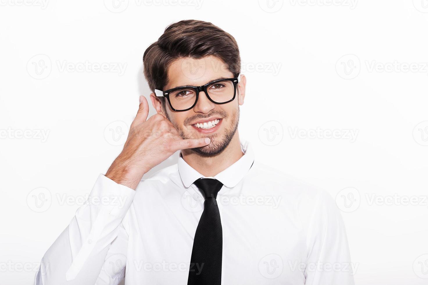 Waiting for your call. Cheerful young man gesturing mobile phone near his face and smiling while standing against white background photo