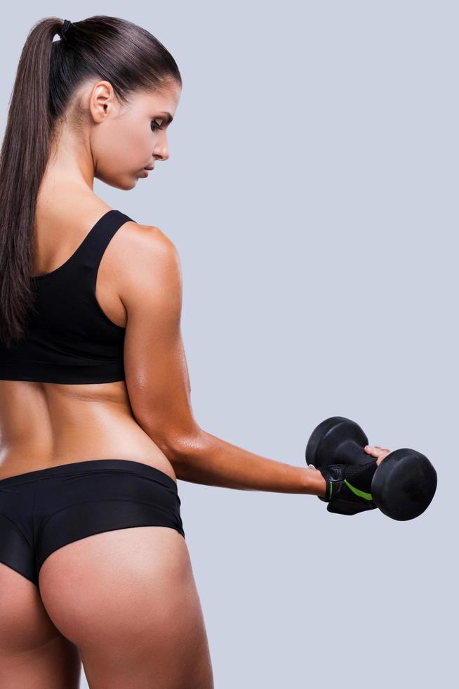 Keeping her body fit. Rear view of young sporty woman with perfect buttocks exercising with dumbbells while standing against grey background photo