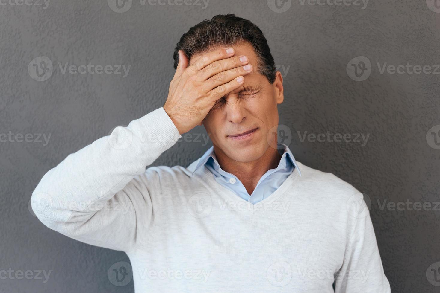 Failed again. Frustrated mature man touching his forehead and keeping eyes closed while standing against grey background photo