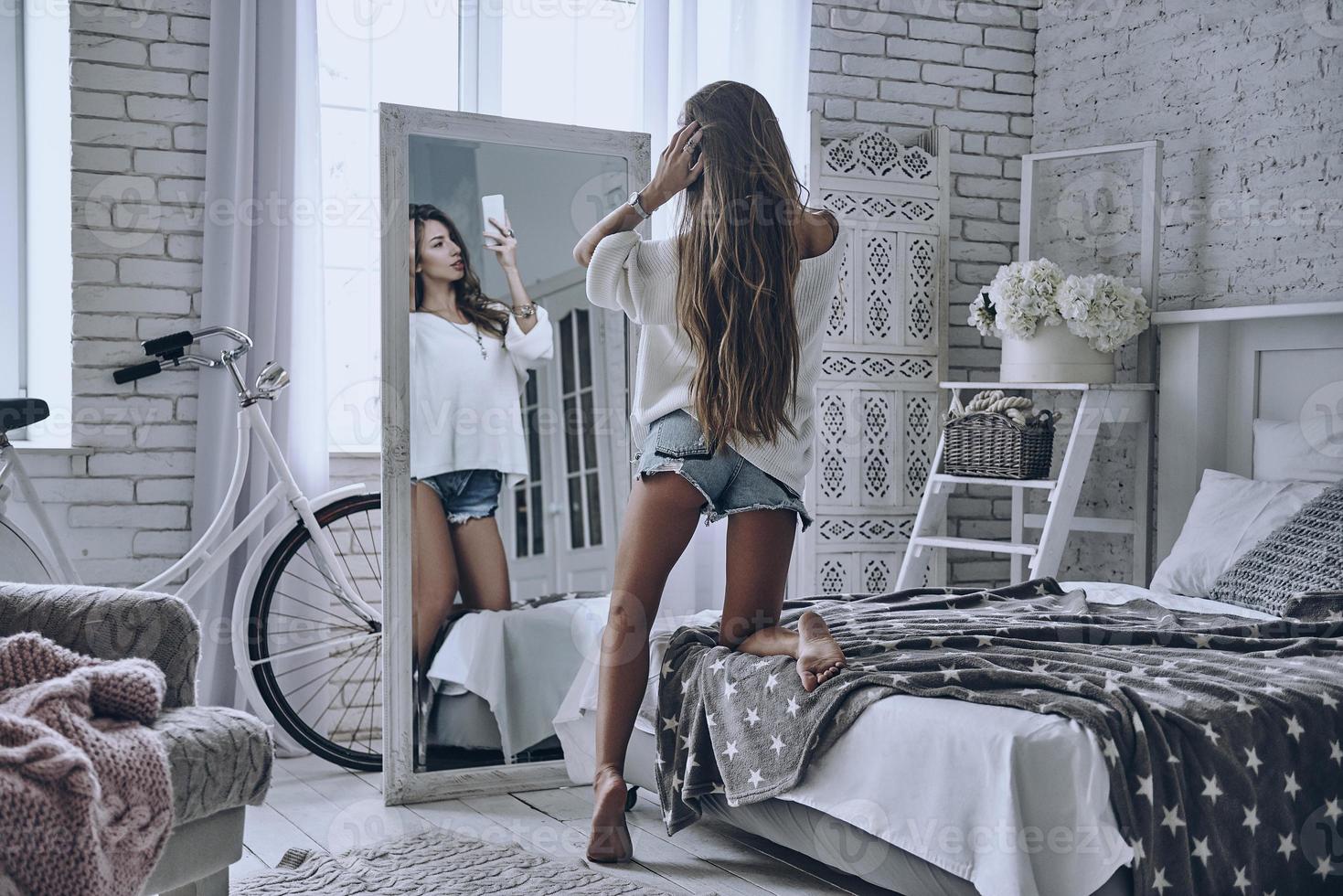 Snapping selfie at home. Rear view of attractive young woman keeping hand in hair and looking in the mirror while taking selfie photo