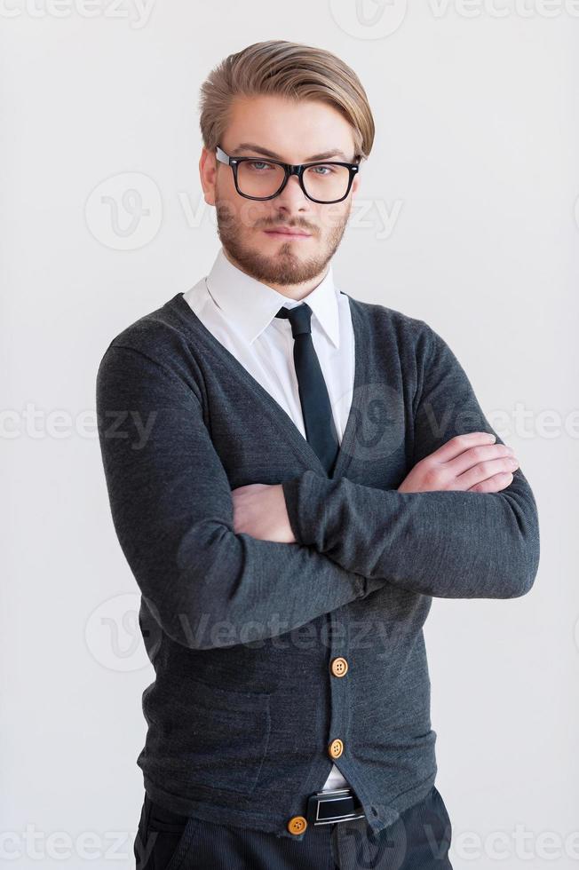 Confident young man. Handsome young man in glasses keeping arms crossed and while standing against grey background photo