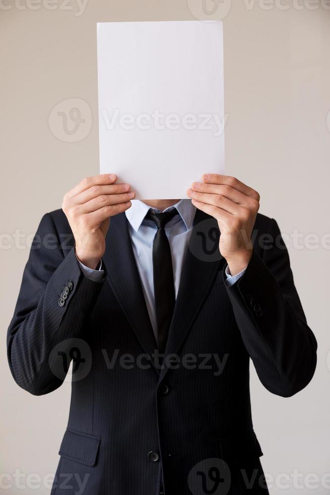 Paperwork in his head. Man in formalwear hiding his face behind blank paper while standing against grey background photo