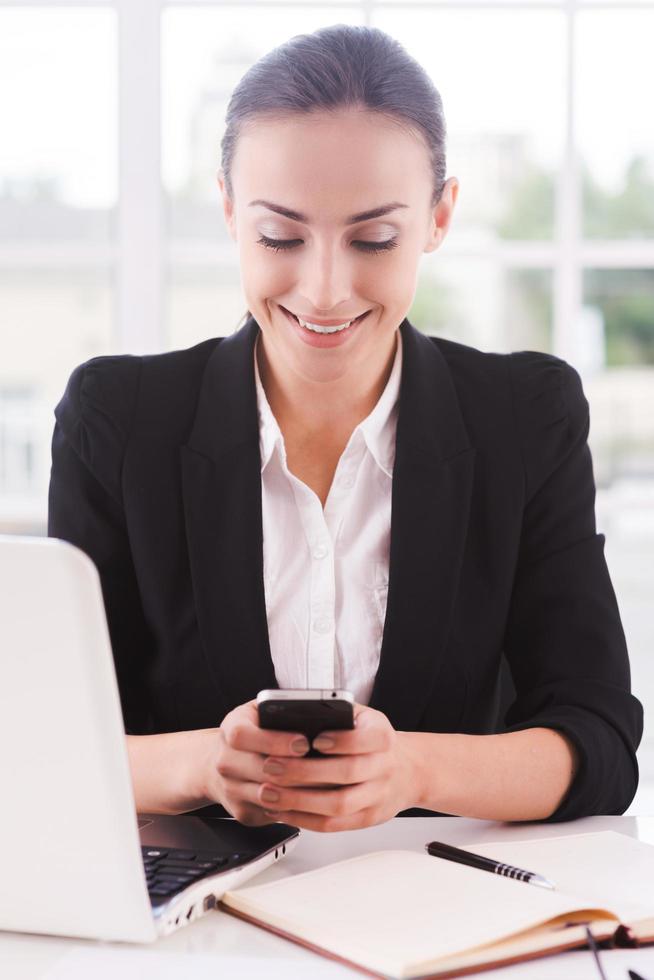 mujer de negocios con teléfono móvil. una joven alegre con ropa formal sosteniendo un teléfono móvil y sonriendo mientras se sienta en su lugar de trabajo foto