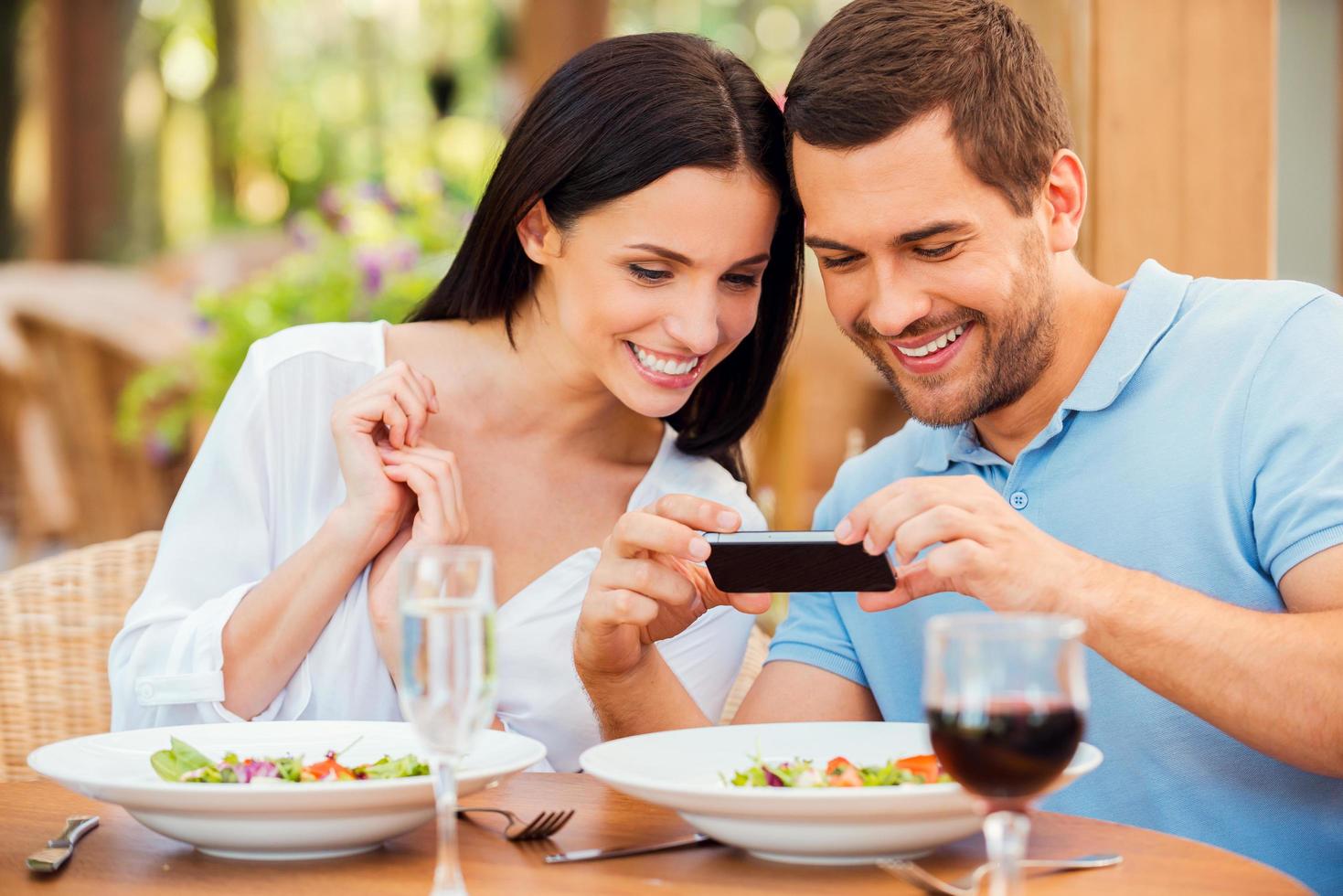 I want to share this picture with friends. Happy young loving couple taking pictures of their food and smiling while relaxing in outdoors restaurant together photo
