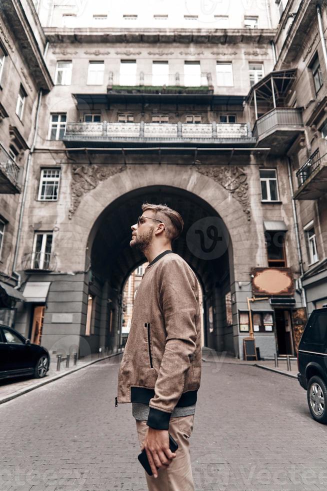 Feeling the city. Handsome young man in casual wear carrying his smart phone and looking away while standing outdoors photo