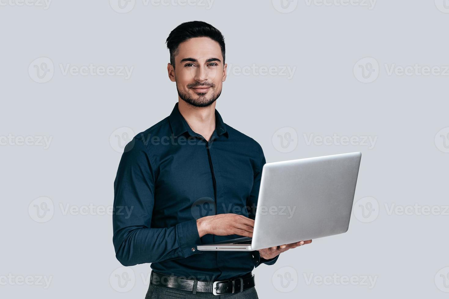 Good looking young man in shirt working on laptop and looking at camera with smile while standing against grey background photo
