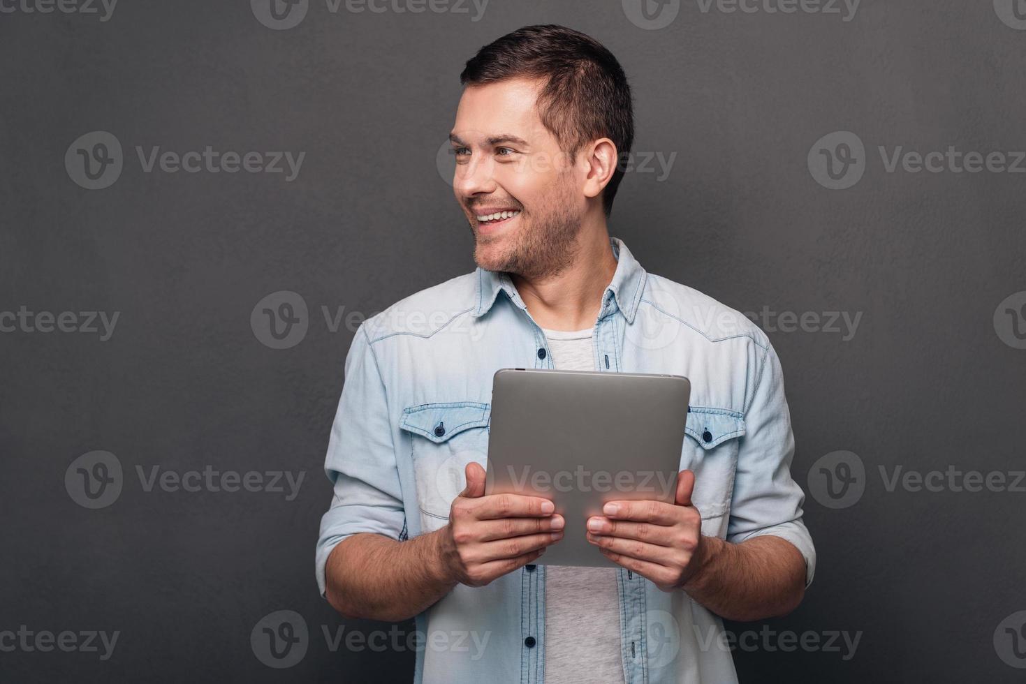I can solve your problem Cheerful young man holding his digital tablet and looking away with smile while standing against grey background photo