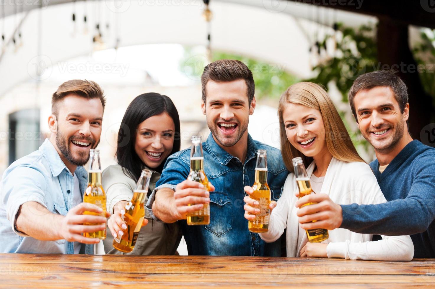 anima a un grupo de amigos de jóvenes felices que se unen entre sí y estiran botellas con cerveza mientras están al aire libre foto