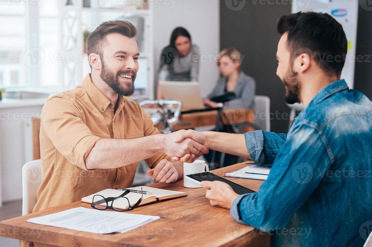 Well done Two confident young men shaking hands and smiling while sitting at the desk in office with two people working in the background photo
