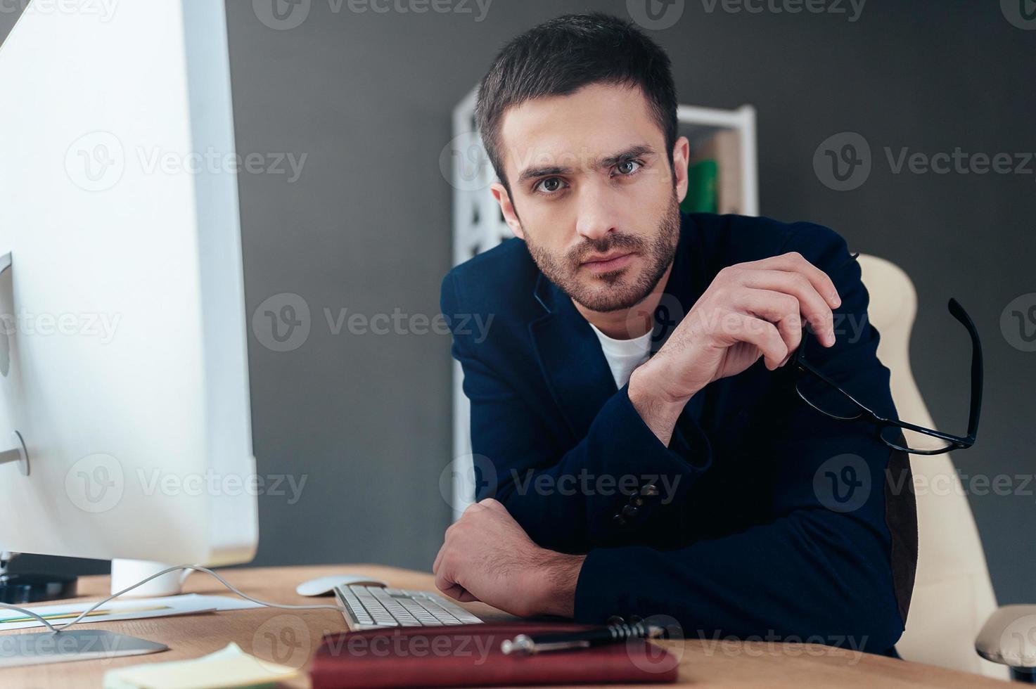 Need an expert advice Confident young man carrying eyeglasses and looking at camera while sitting at his working place in office photo